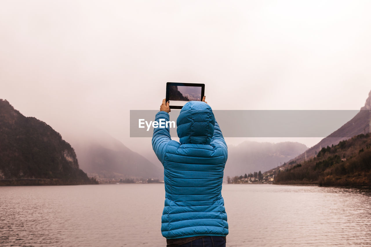 Rear view of woman photographing with digital tablet by lake during foggy weather