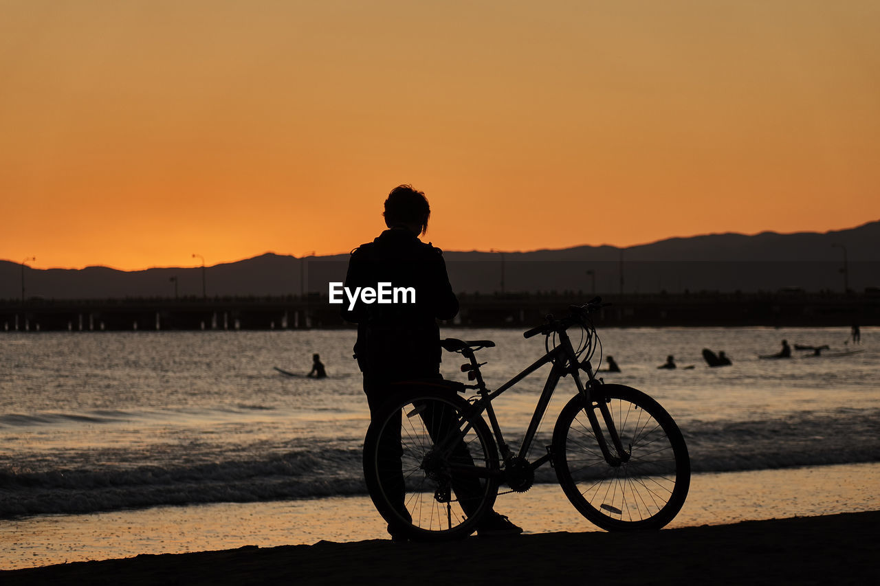 Silhouette woman with bicycle on beach against sky during sunset