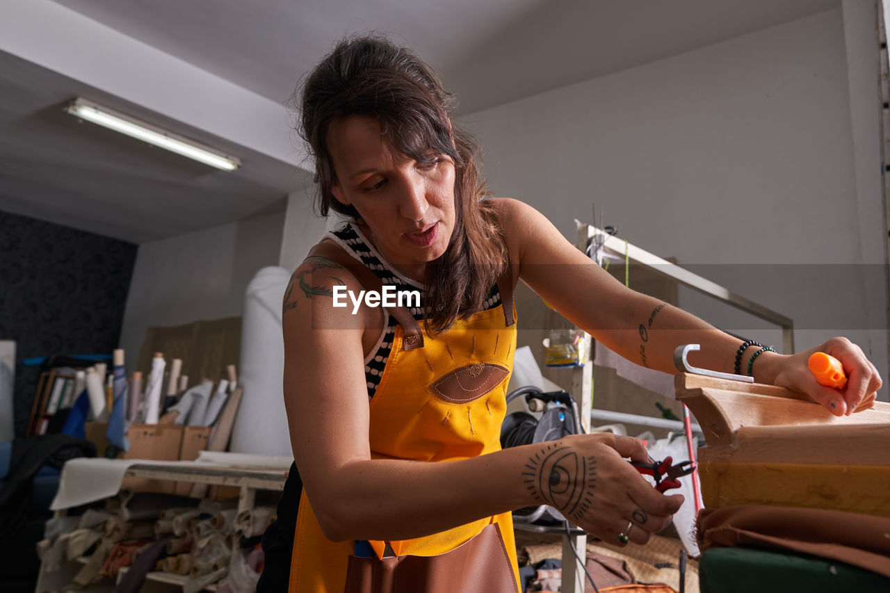 Concentrated adult tattooed female artisan with dark hair in apron using pliers during repairing process at wooden furniture in workshop