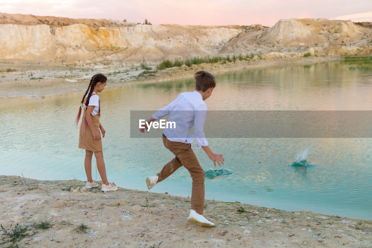 Teenagers 8-9 years old throw stones into water from shore, outdoor entertainment together.