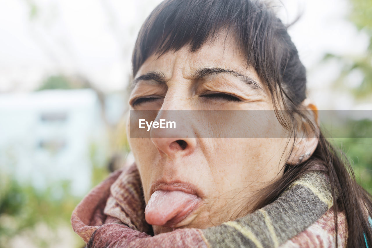 Portrait of woman eyes closed while sticking out tongue outdoors