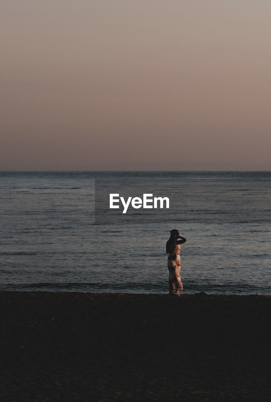 rear view of woman standing at beach against clear sky during sunset