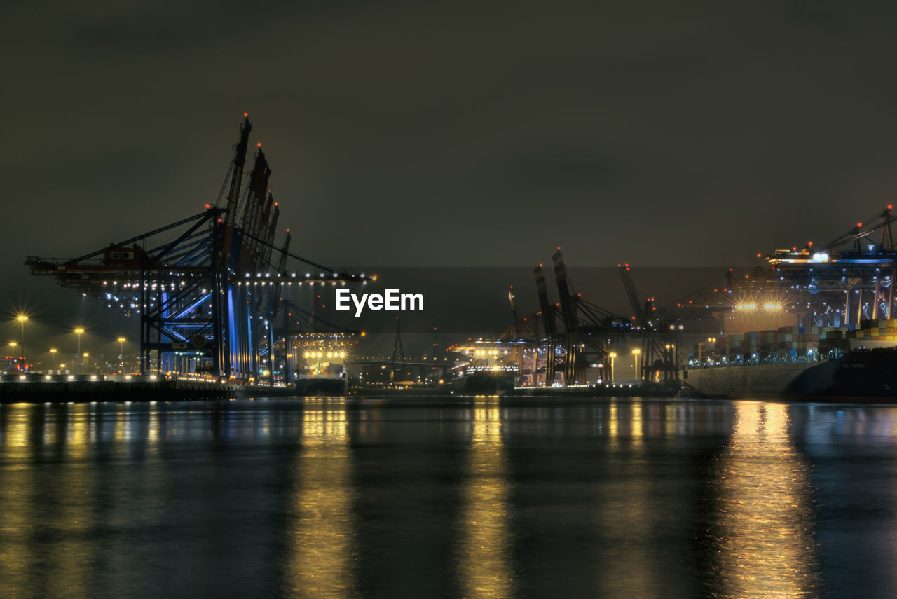 Illuminated cranes over sea against sky at night