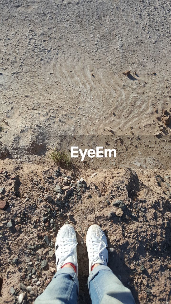 low section, human leg, shoe, land, personal perspective, one person, high angle view, standing, lifestyles, day, nature, sand, leisure activity, beach, men, soil, outdoors, geology, limb, rock, sunlight, human limb, jeans, casual clothing, sea, human foot, footwear, water