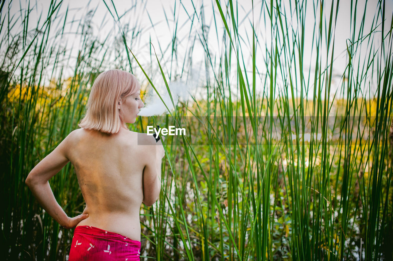 Rear view of shirtless young woman smoking while standing on grassy field