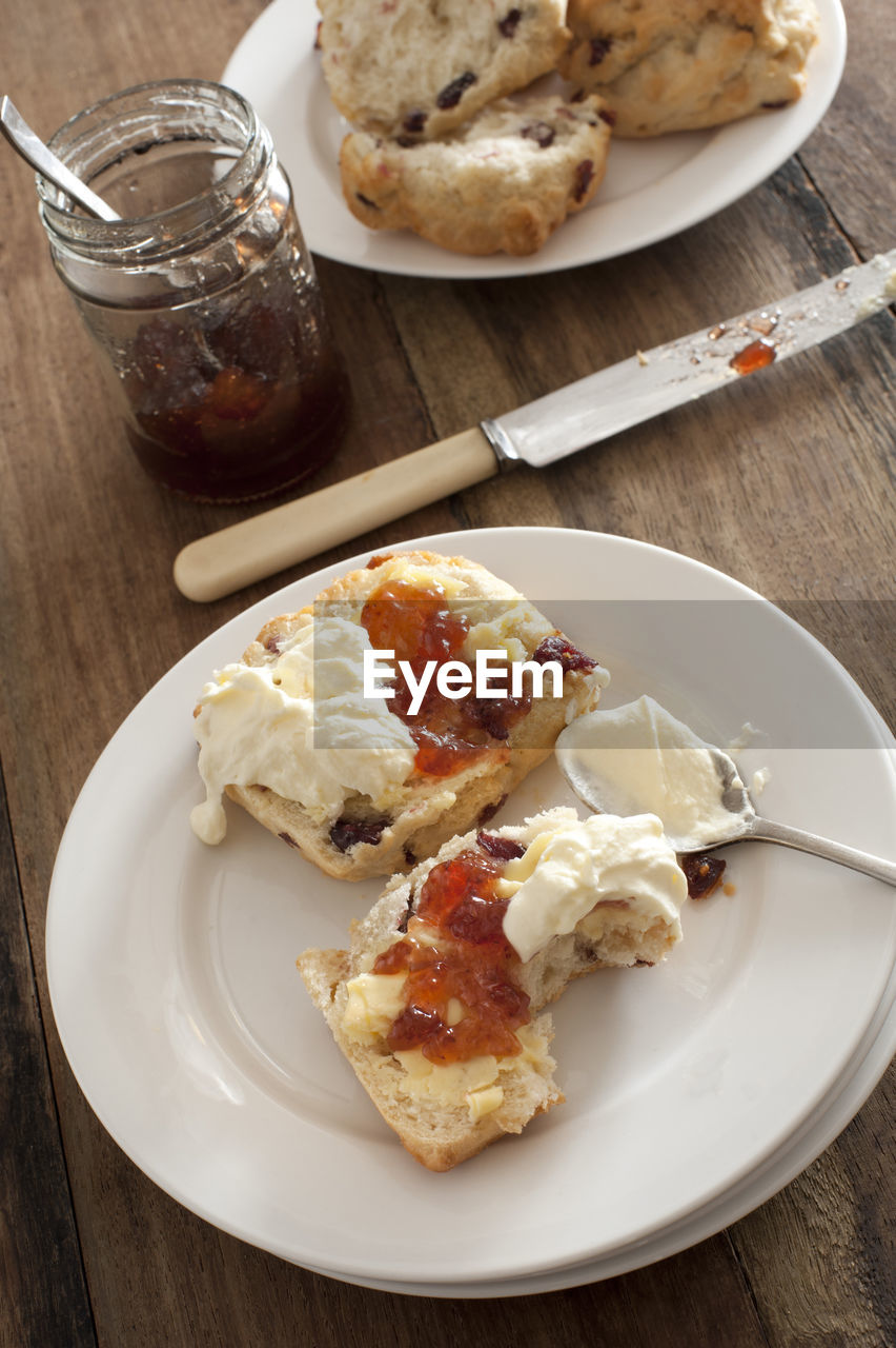 Tasty snack of a raisin scone or rock cake, fruit jam and whipped cream on a white plate with a bite