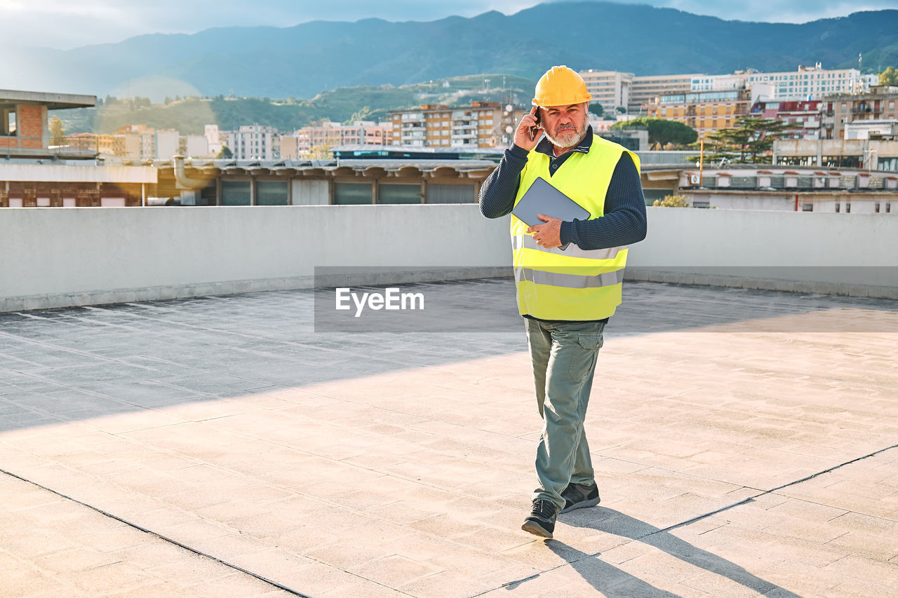 Middle aged supervisor in hardhat and safety vest talking worried on smartphone on building site. 