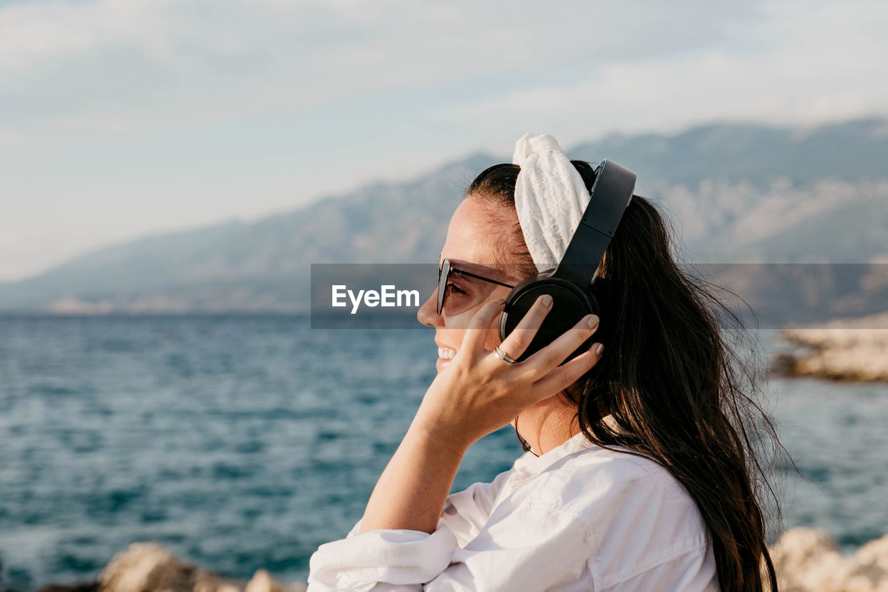 Side view of young woman in white shirt listening to music on headphones. summer, beach, lifestyle.