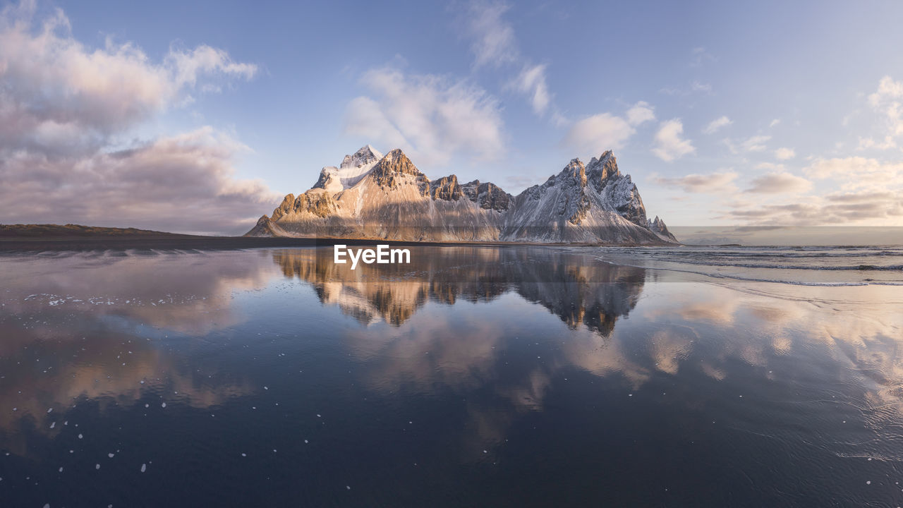 Spectacular nordic scenery of calm frozen lake near rocky vestrahorn mountain with snowy peaks during colorful sunset at stockness beach, iceland