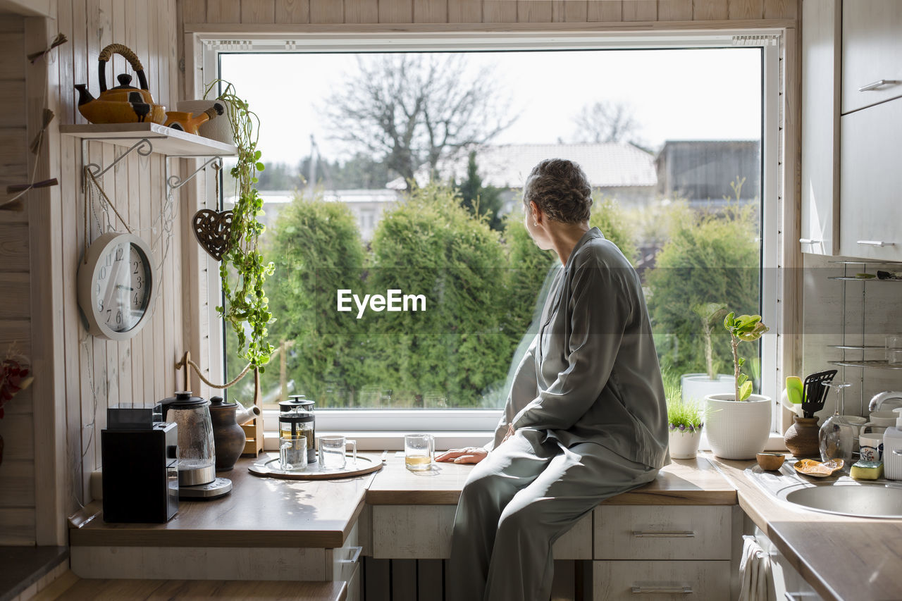 Mature woman sitting on kitchen counter at home looking out of window