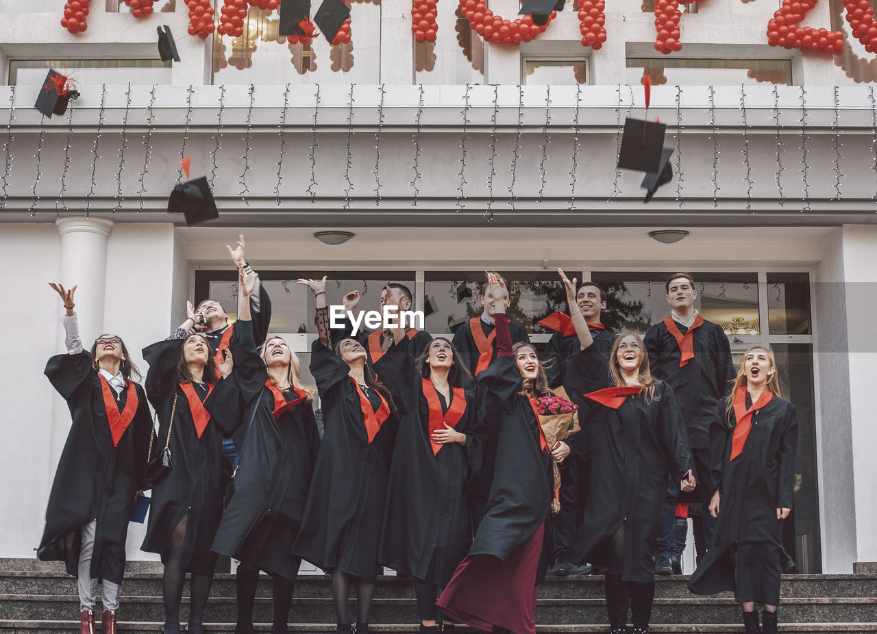 Low angle view of happy students wearing graduation gowns standing on steps by building