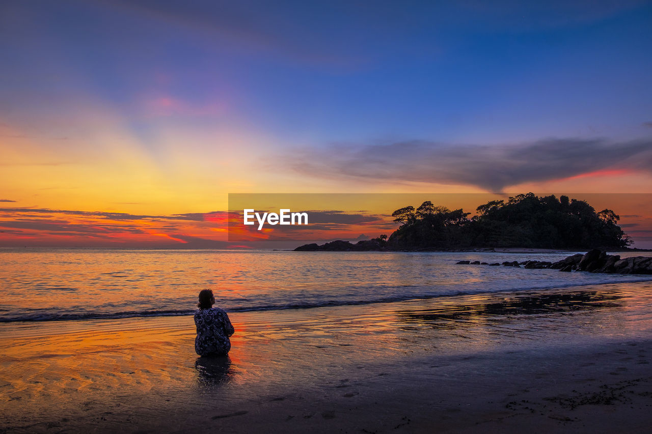 REAR VIEW OF PERSON STANDING ON BEACH DURING SUNSET