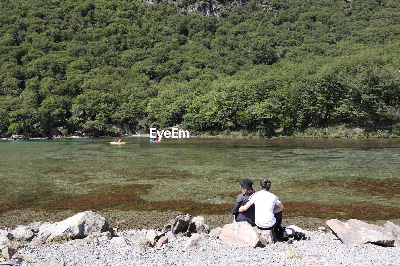 tree, plant, water, river, nature, leisure activity, sitting, men, beauty in nature, day, lifestyles, rock, wilderness, scenics - nature, green, rear view, growth, land, tranquility, adult, non-urban scene, outdoors, one person, tranquil scene, mountain, casual clothing, full length, forest, beach, sunlight, women, idyllic, relaxation, remote