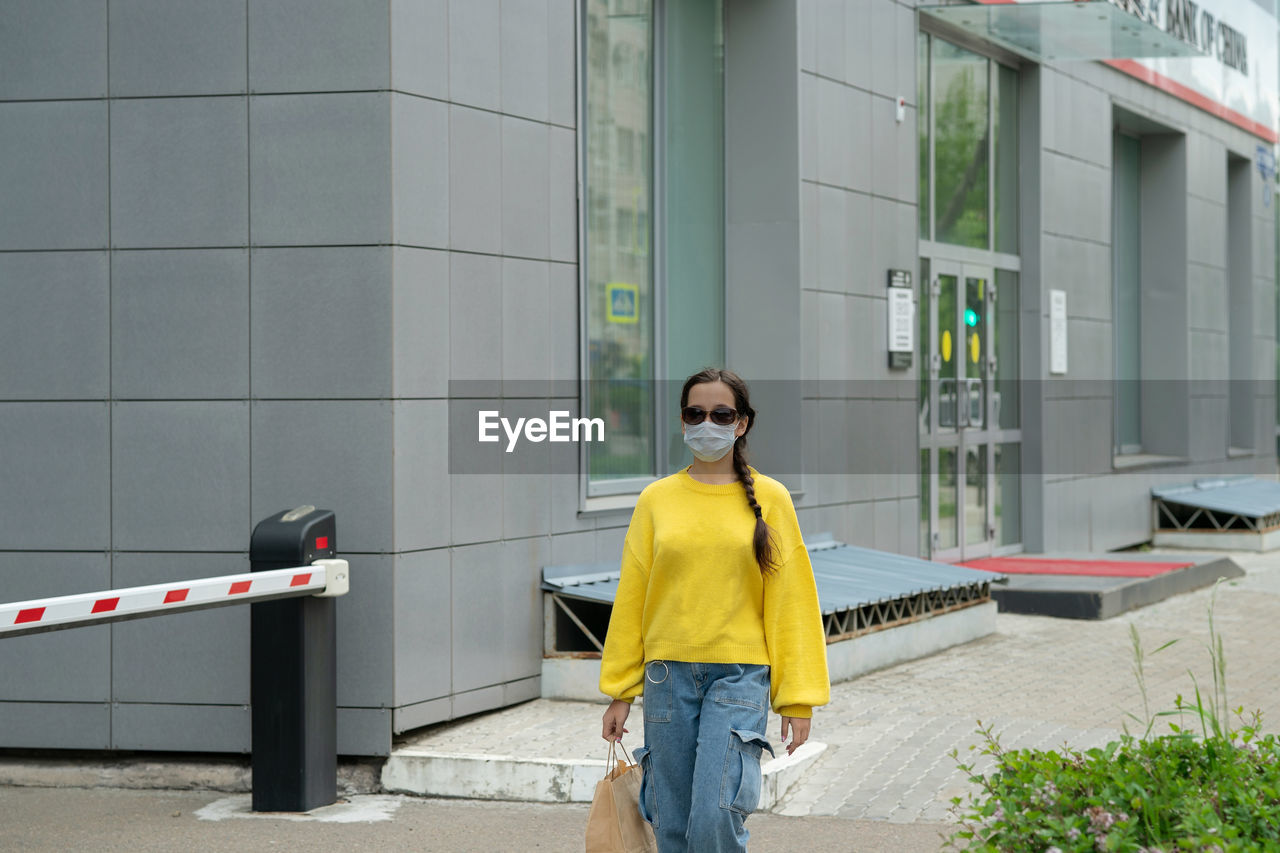 Girl in a yellow sweater and jeans with a mask and sunglasses on her face walks past a bank of china