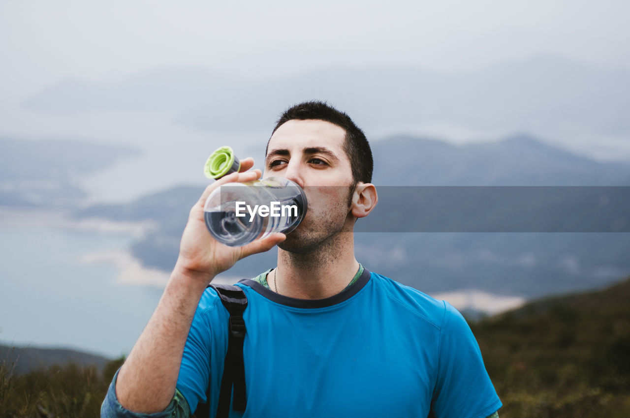 Male hiker drinking water while standing on mountain against sky
