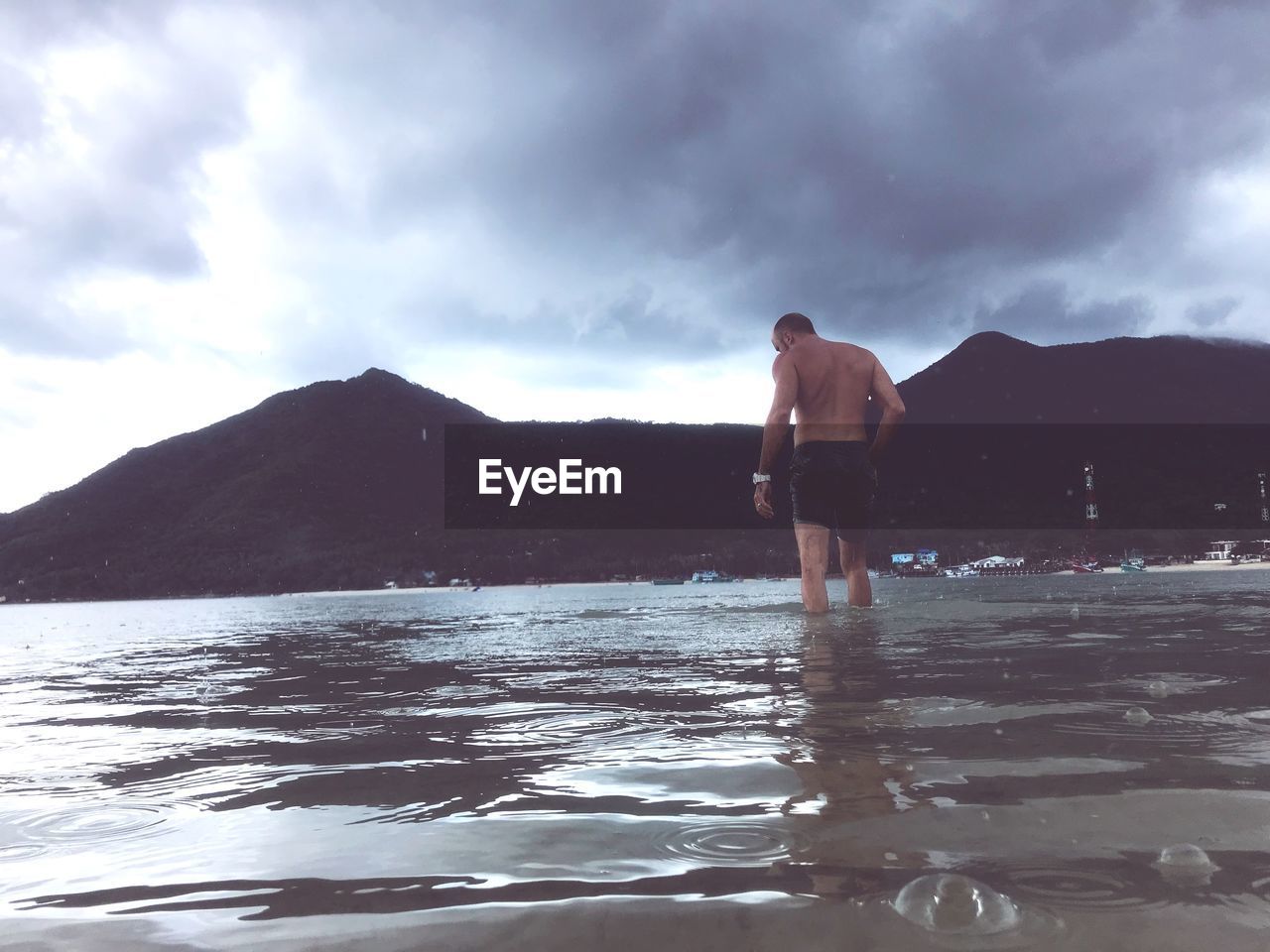 REAR VIEW OF SHIRTLESS MAN STANDING IN LAKE AGAINST SKY