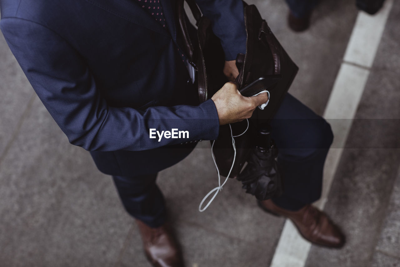 Low section of businessman with open bag holding smart phone and in-ear headphones in city