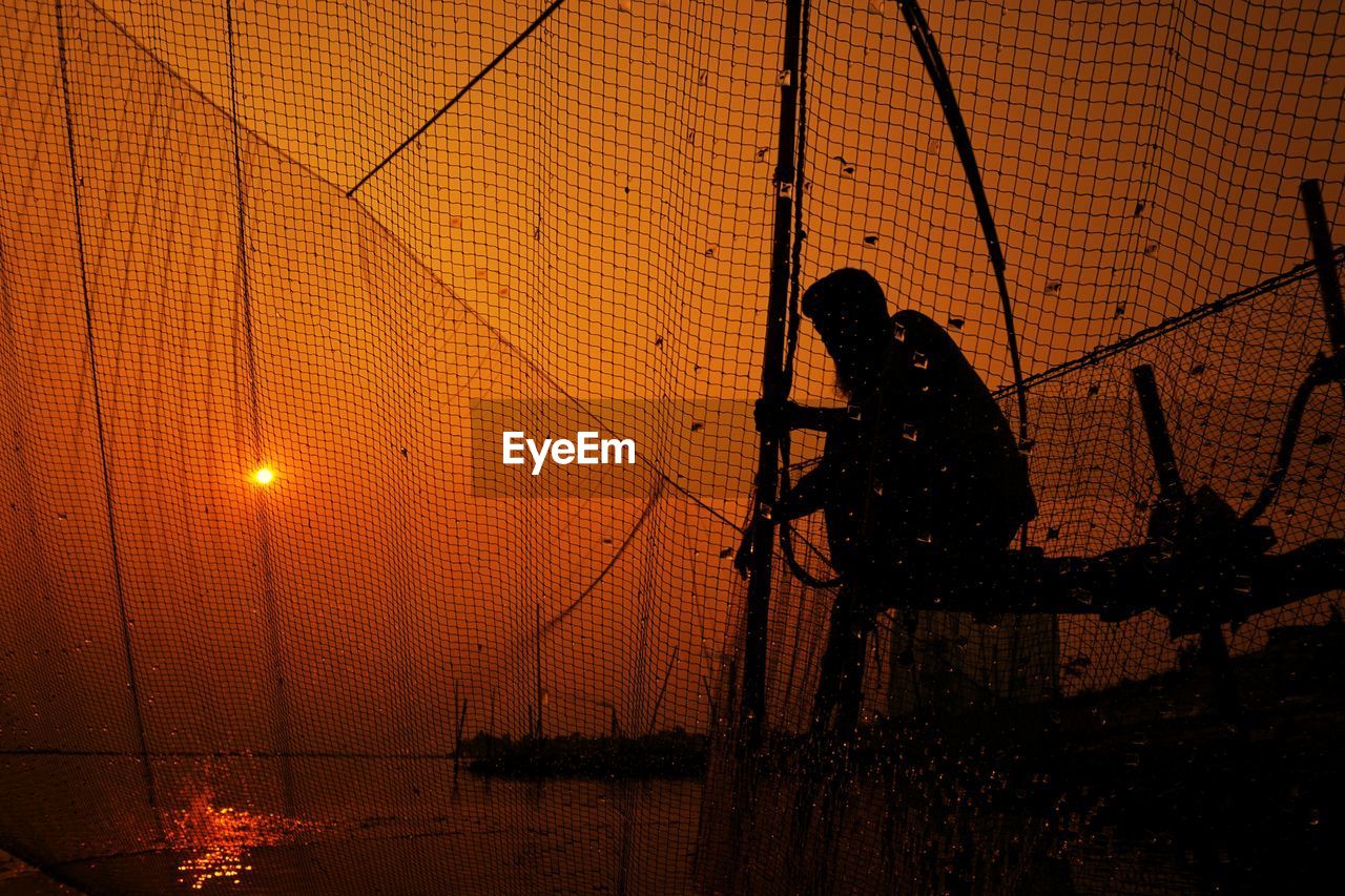 Silhouette man against orange sky seen through fence during sunset