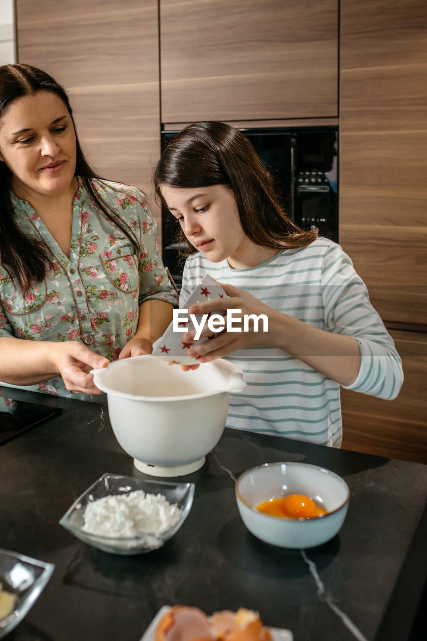 Young girl pouring flour into bowl while baking with mother in kitchen