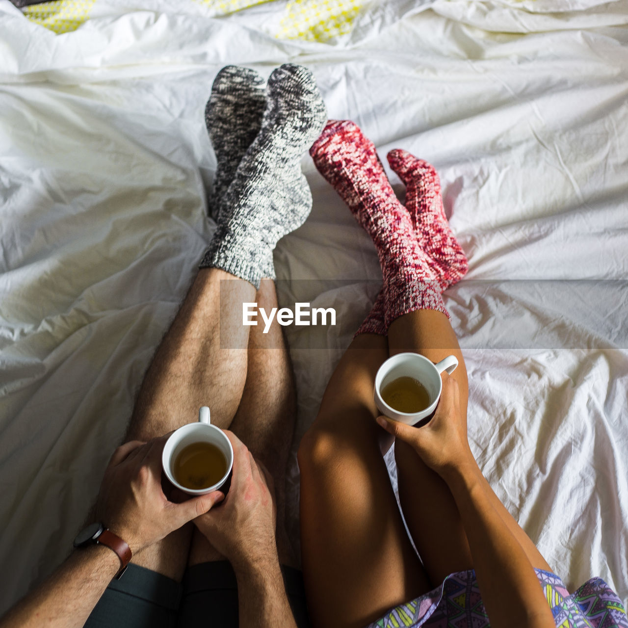 Low section of couple holding tea cup while wearing socks on bed at home