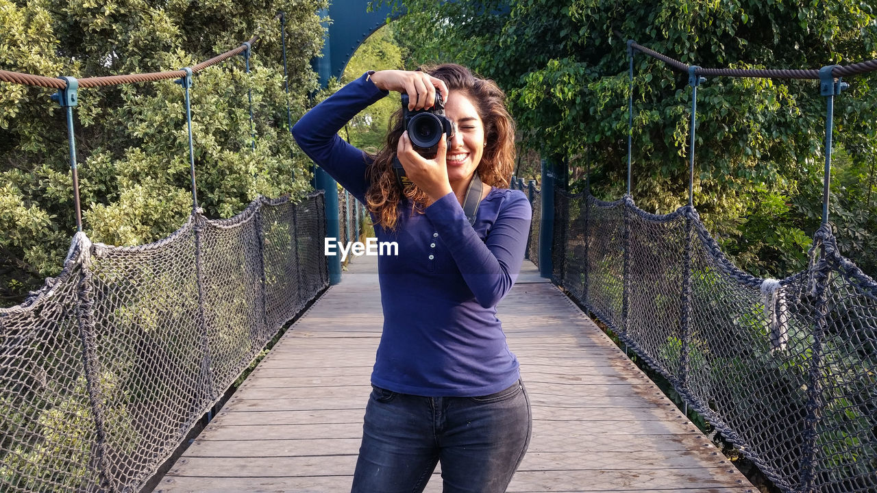 Smiling young woman photographing with camera while standing on footbridge in park