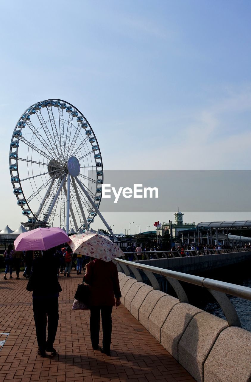 Rear view of women with umbrellas while walking by ferris wheel against sky