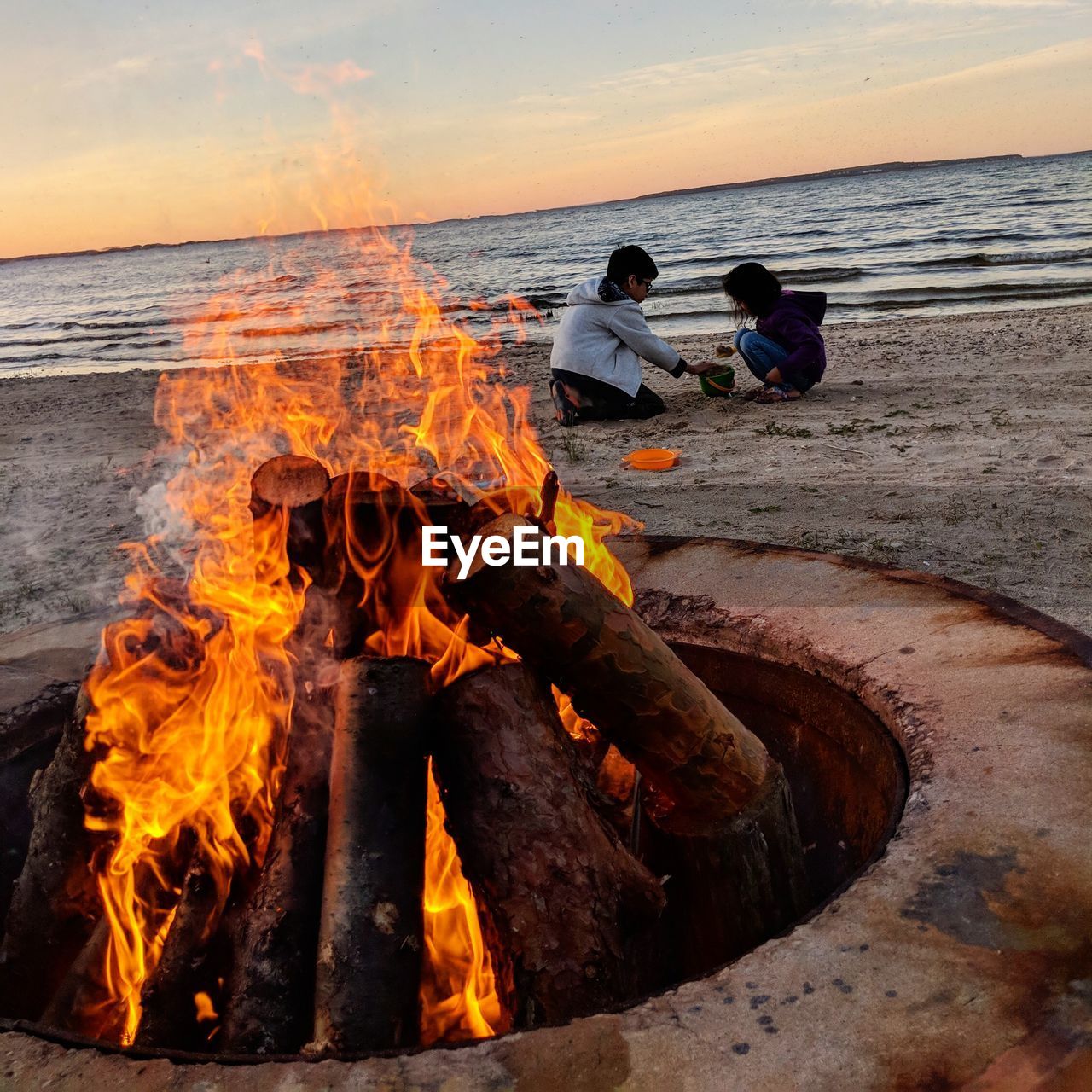 Campfire at beach with siblings playing on sand against sky during sunset