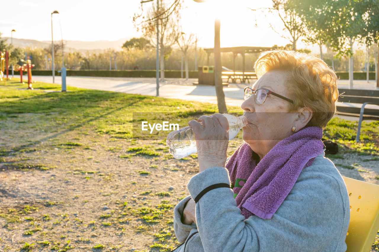 Elderly woman drinks water from the bottle after exercising in a park at sunset