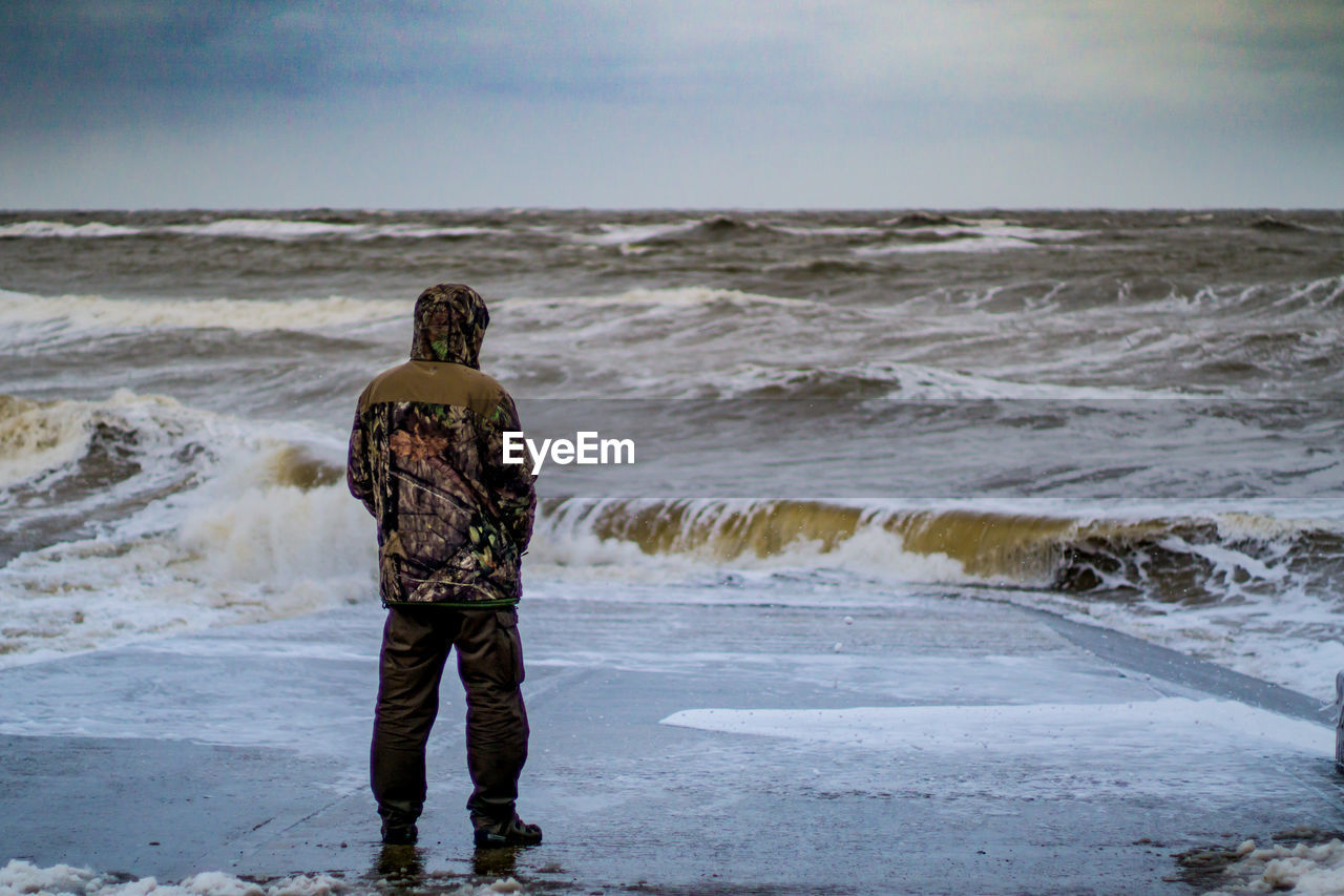 Rear view of man standing on shore at beach against sky