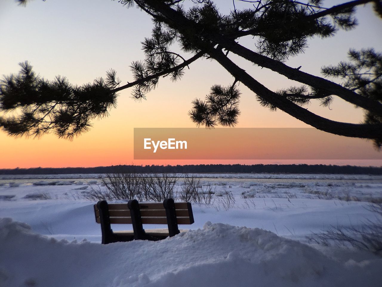 Bench on snow covered field against clear orange sky