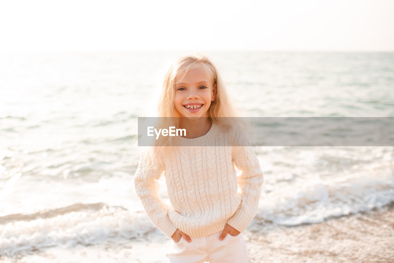 Happy smiling child girl 7-8 year old wear white knitted sweater and pants posing over nature sea