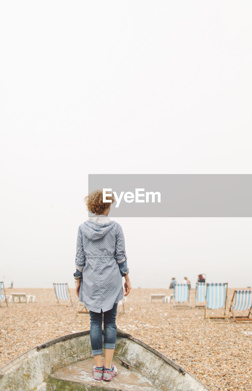 Rear view of woman standing in old boat at beach against clear sky