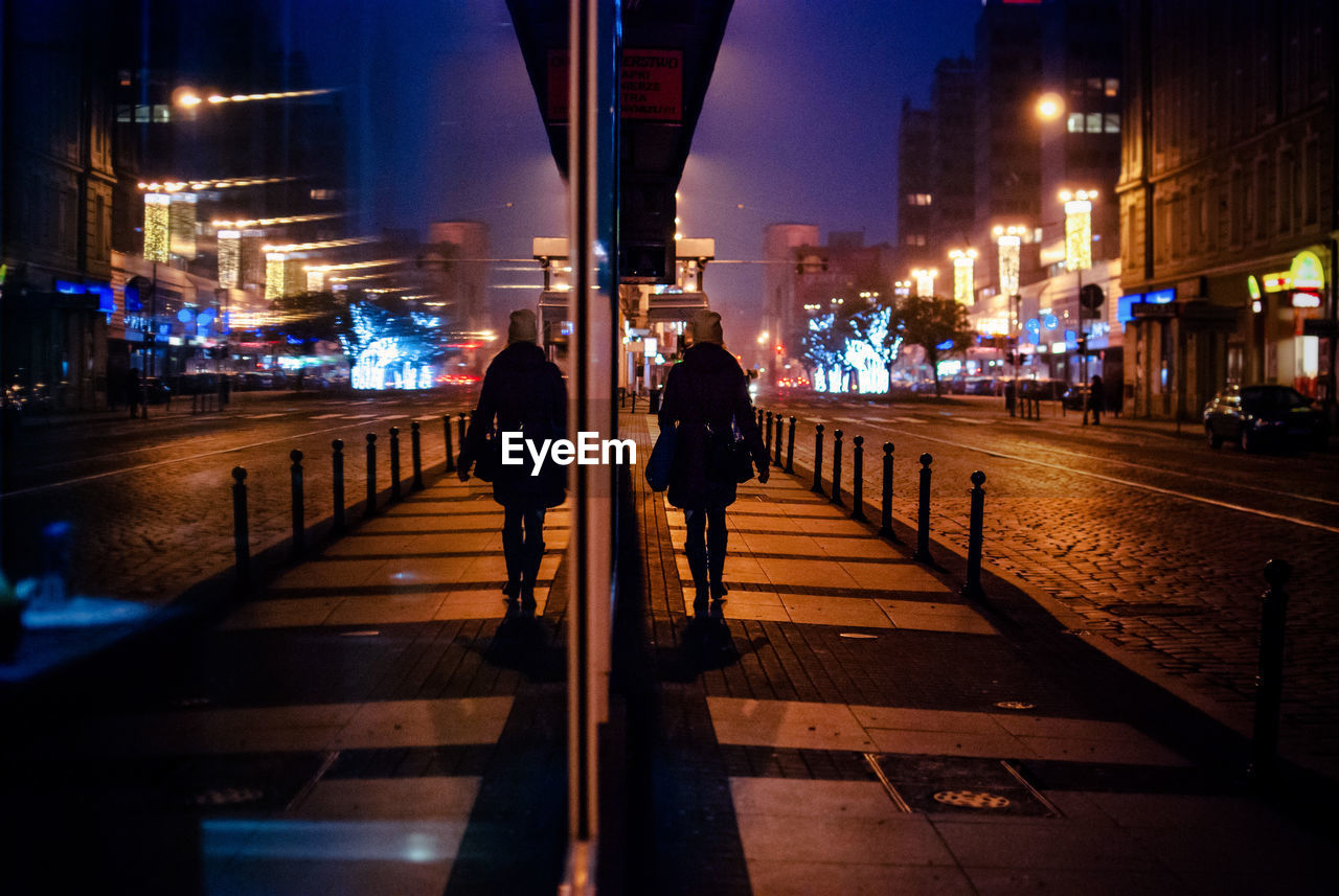 Rear view of woman walking on sidewalk reflecting on window at night