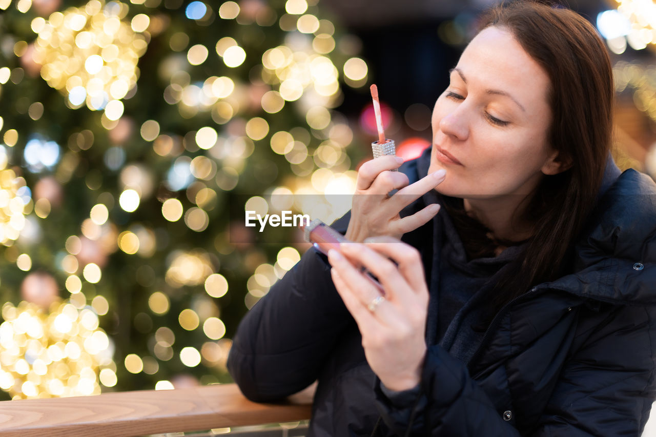 Woman paints her lips on a background with christmas tree lights bokeh