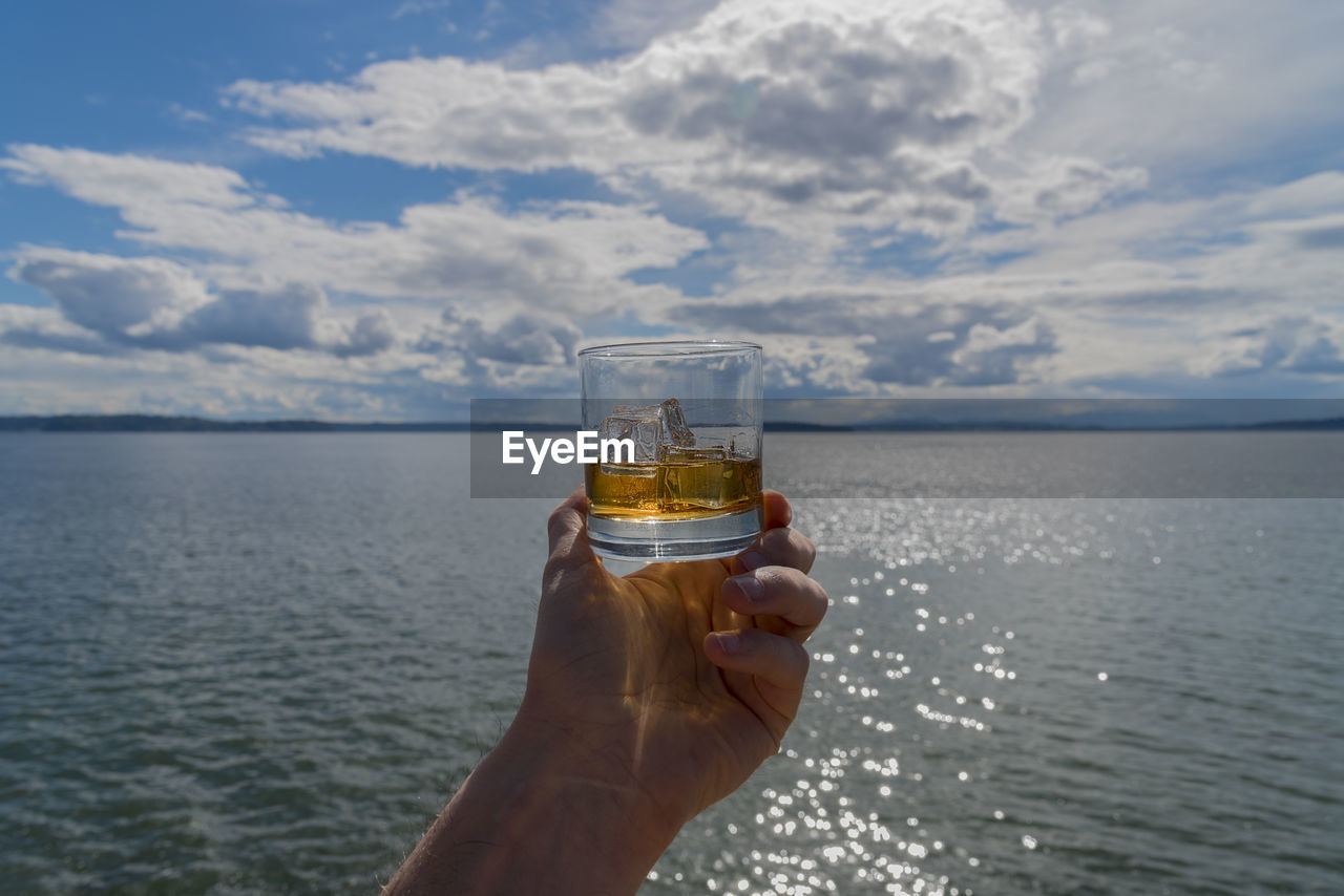 Cropped hand of person holding whisky glass against sea and cloudy sky