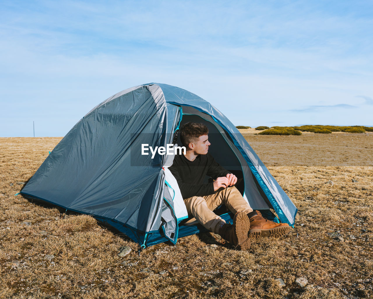 Full body of trendy young male traveler in stylish outfit sitting in camping tent while resting after trekking