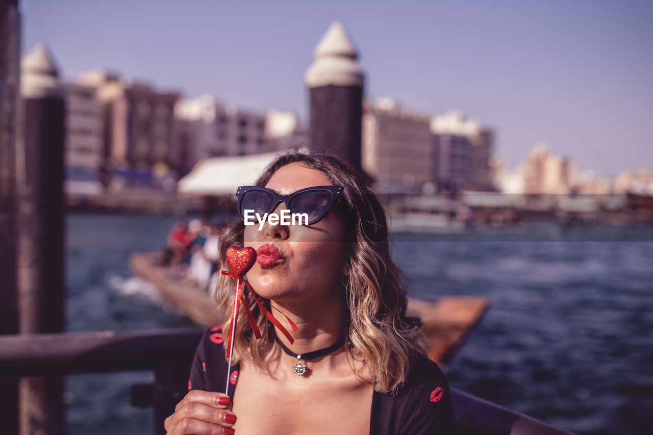 Woman wearing sunglasses puckering at heart shape candy against river in city