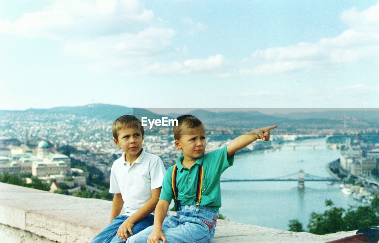 Boy sitting with brother pointing on bridge over river amidst city