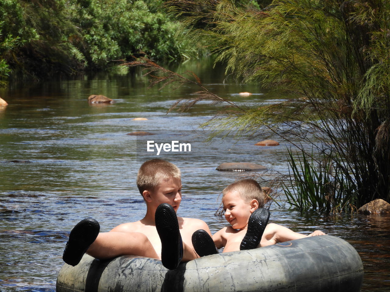 Shirtless brothers floating on inflatable ring over river against trees