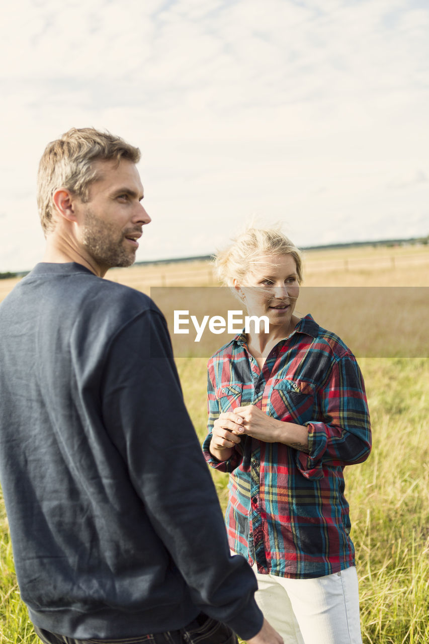 Man and woman looking away while standing on grassy field against sky