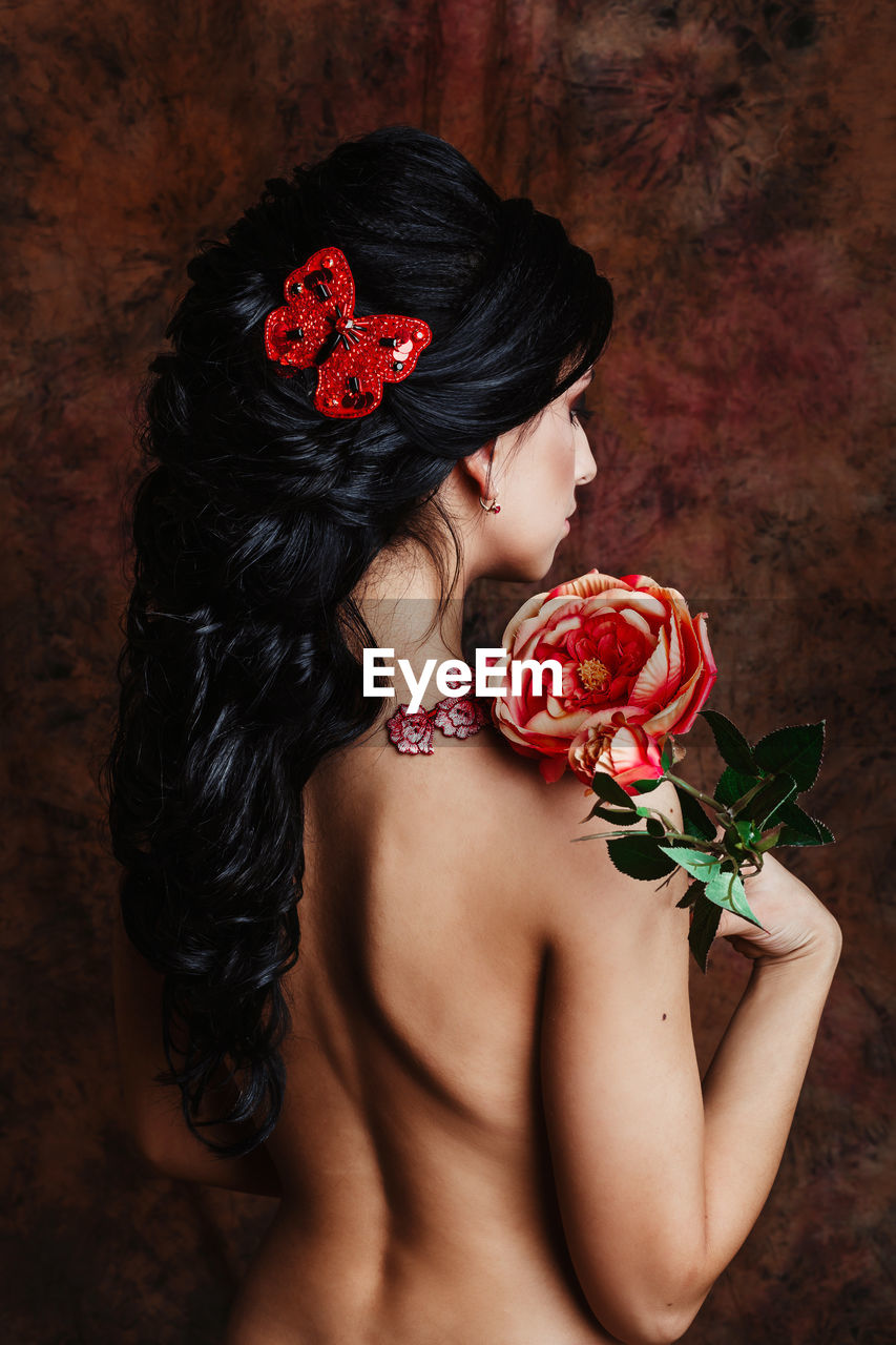 Rear view of shirtless young woman holding flower standing against wall