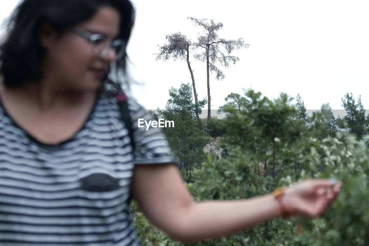 plant, one person, nature, casual clothing, adult, tree, looking, young adult, day, outdoors, waist up, person, glasses, growth, sky, portrait, standing, holding, front view, striped, eyeglasses, grass, selective focus, t-shirt, flower, leisure activity, hairstyle, emotion, headshot