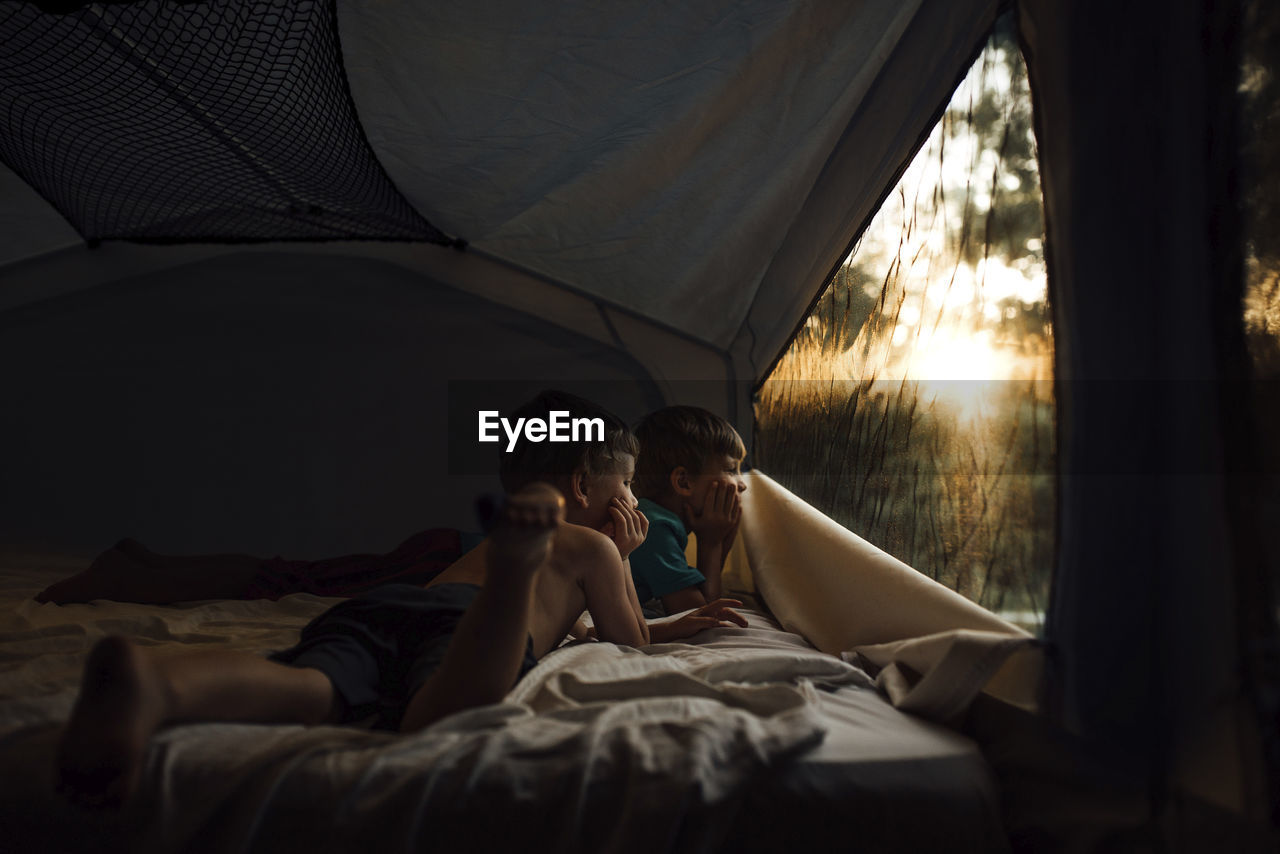 Thoughtful brothers looking through window while lying in tent