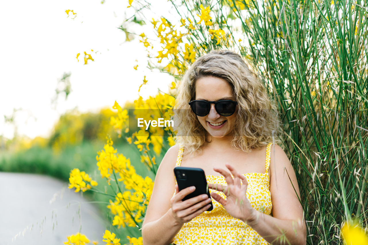 YOUNG WOMAN USING SMART PHONE WHILE STANDING ON YELLOW CAMERA