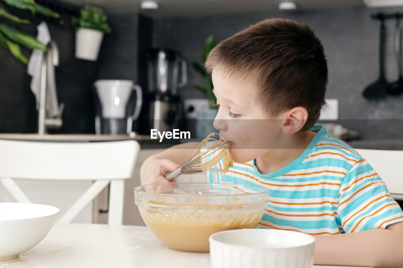 Cute boy licks a whisk with cake dough in the kitchen at home. time with children at home. 