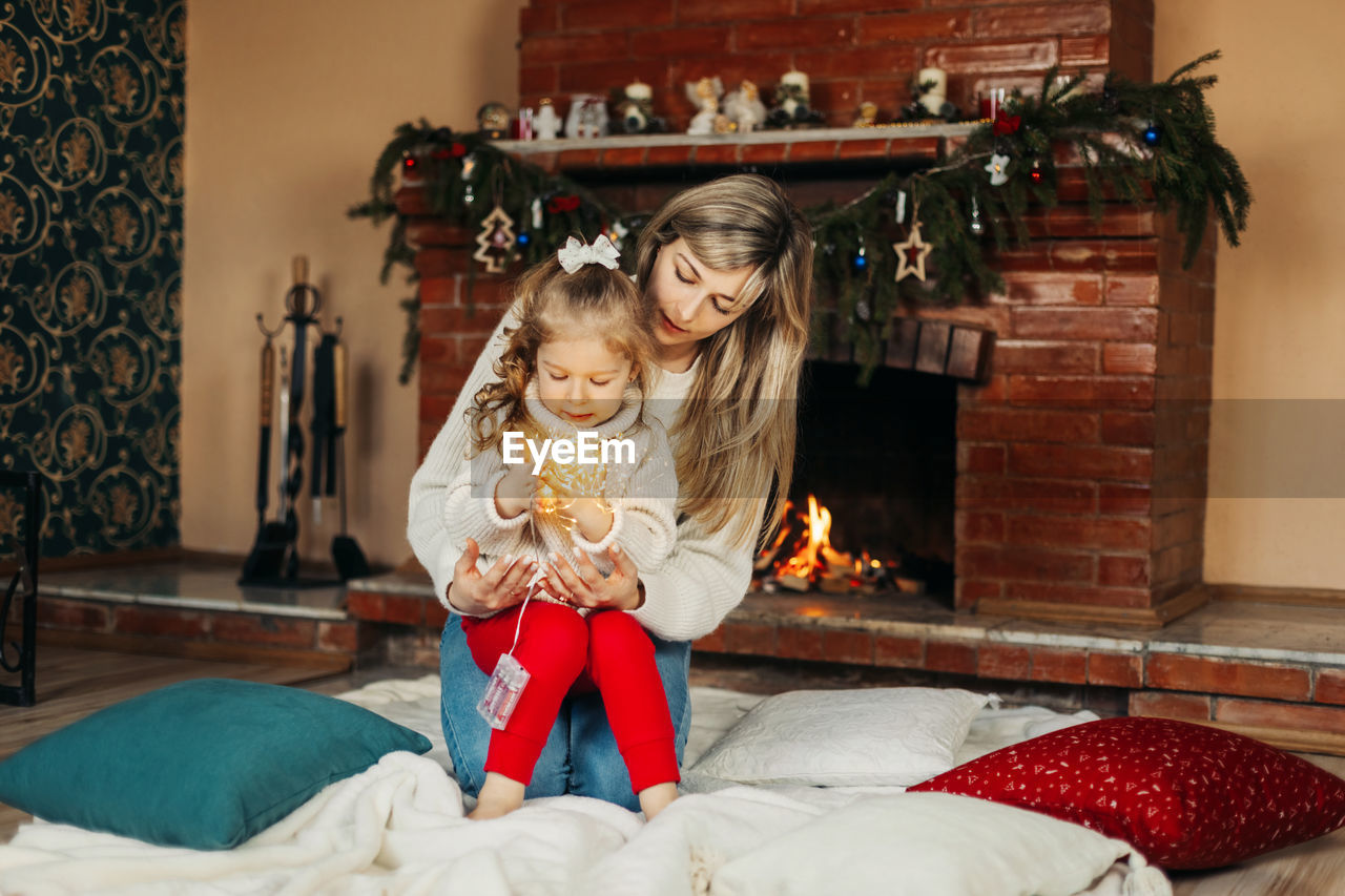 Mom and daughter are playing with christmas garlands on the floor by the fireplace