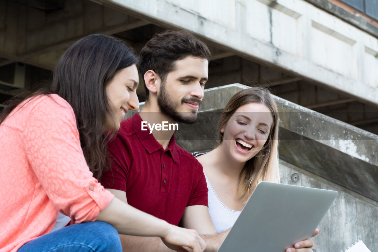 SMILING YOUNG WOMAN USING MOBILE PHONE WHILE SITTING ON LAPTOP IN SMART