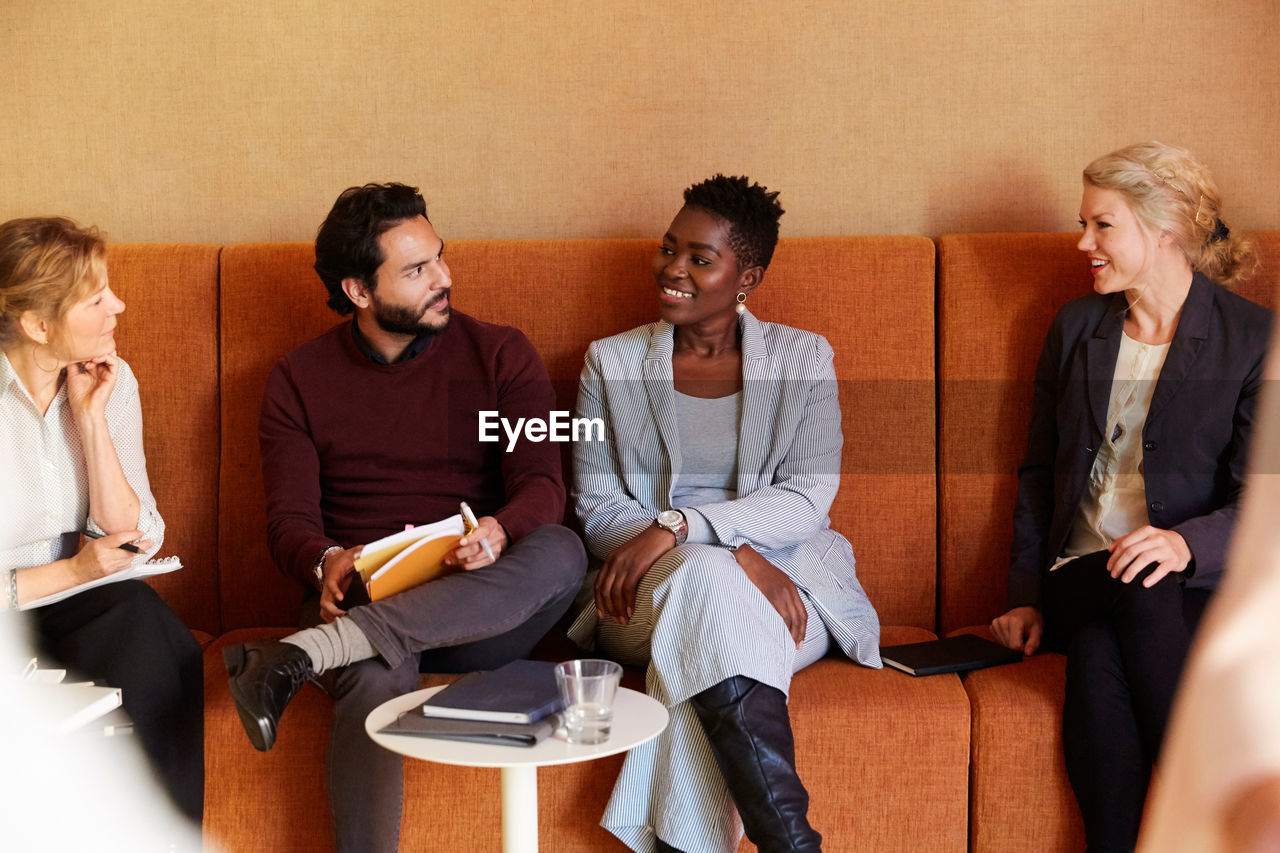 Smiling male and female entrepreneurs sitting with coworkers on sofa at workplace