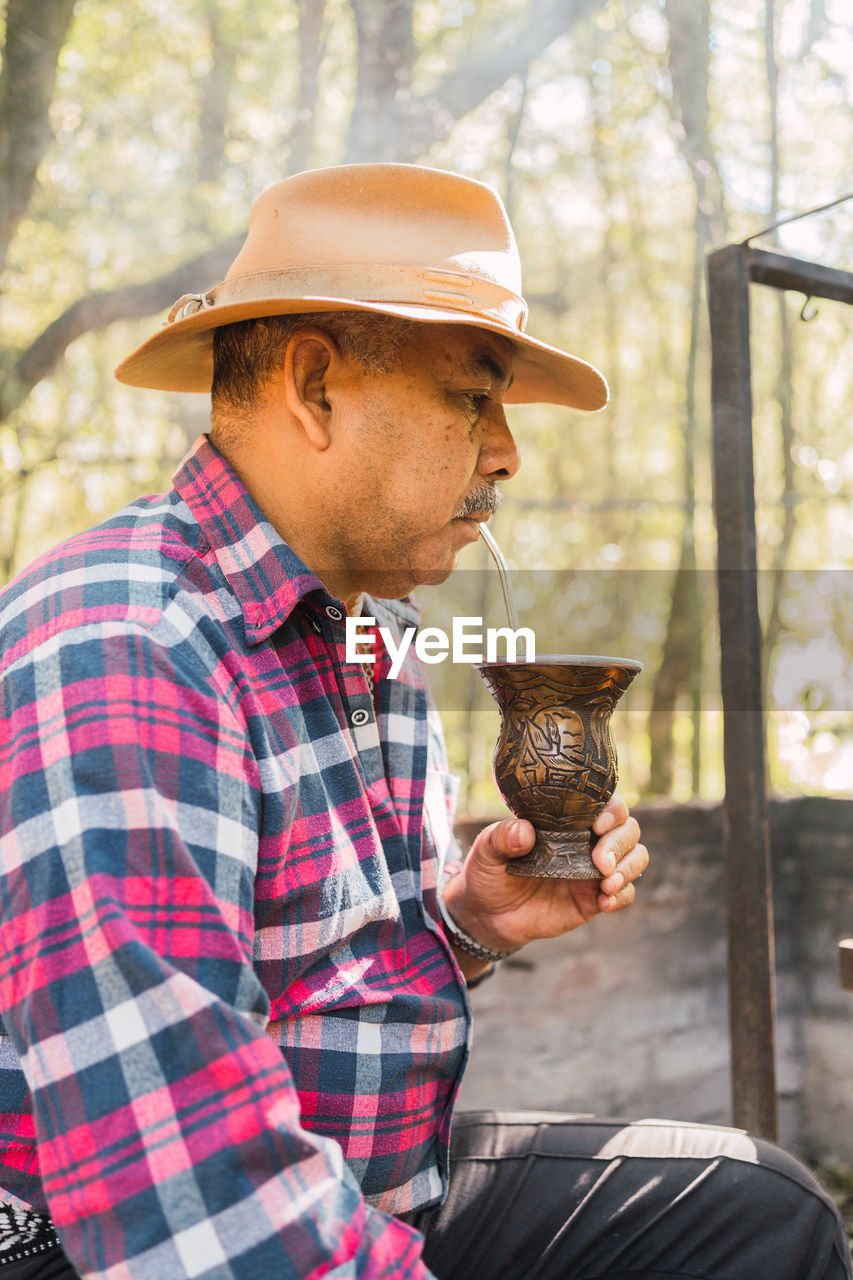 Side view of argentinian male drinking mate from calabash gourd near kettle against rack above burning fire in countryside