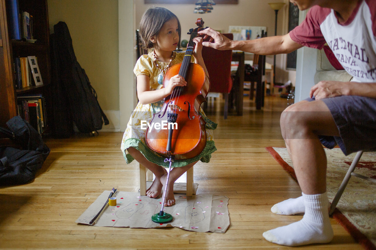 A father leans in to help a little girl practice playing a cello